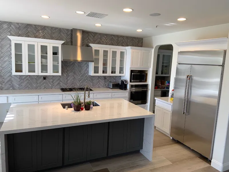 Kitchen with a white quartz waterfall island on dark base cabinets, glass front uppers and a gray herringbone backsplash